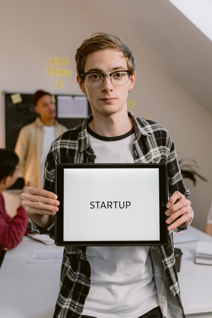 A young professional stands in a startup office holding a digital tablet displaying the word STARTUP.